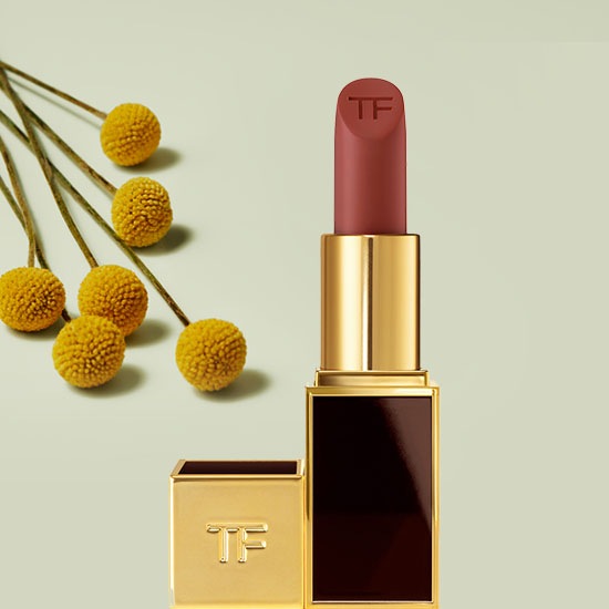 Szminka Tom Ford w złotym opakowaniu z brązowym sztyftem, obok której leżą żółte kwiaty.