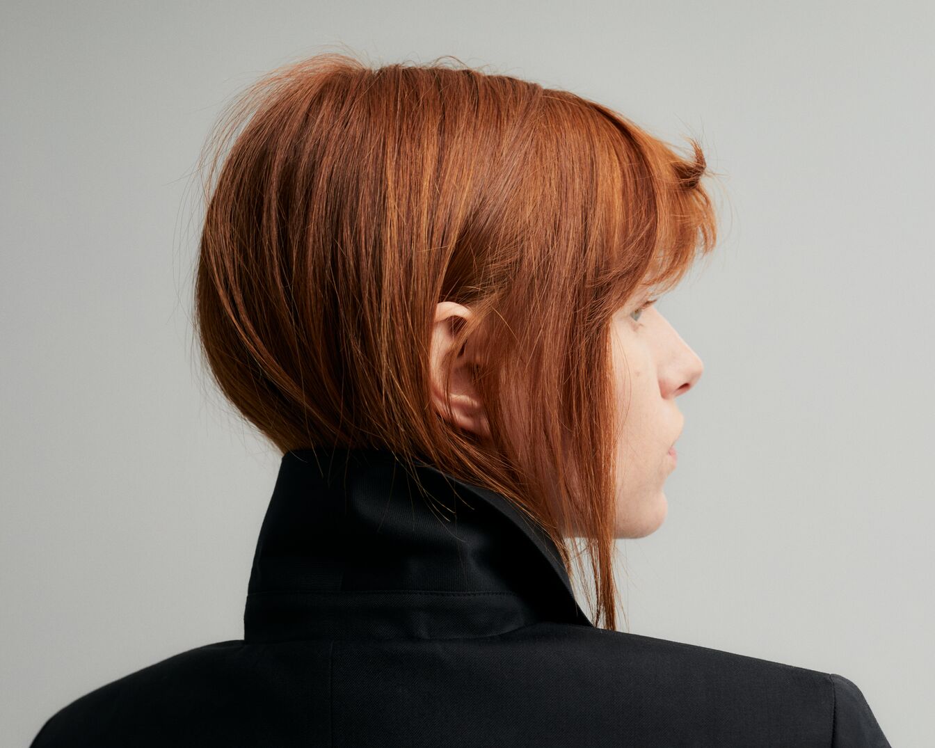 Vista lateral de una mujer con cabello corto y rojizo, vestida con una chaqueta negra de cuello alto, sobre un fondo gris claro.