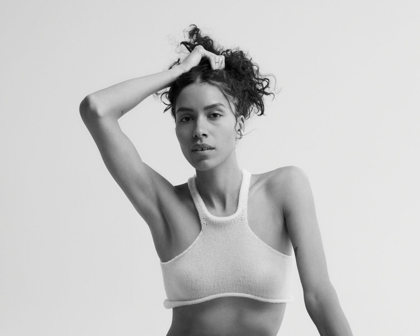 En una foto en blanco y negro, una modelo de piel morena y cabello rizado posa con un top tejido blanco, levantando una mano para sujetar su cabello.