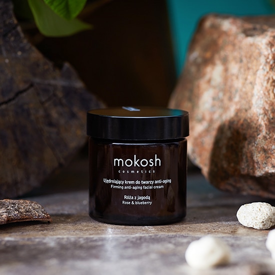 Słoik brązowego kremu do twarzy Mokosh Cosmetics z czarną zakrętką, otoczony kamieniami i zielonymi liśćmi. Krem jest oznaczony jako ujędrniający, przeciwstarzeniowy krem do twarzy z różą i jagodą.