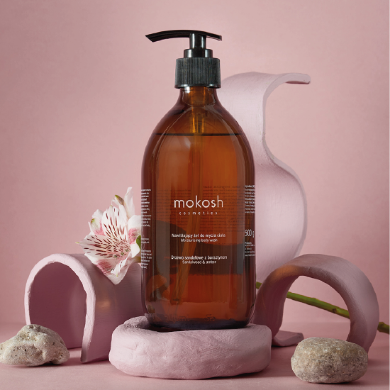 Butelka z brązowego szkła z dozownikiem zawiera nawilżający żel do mycia ciała Mokosh o zapachu drzewa sandałowego i bursztynu. Produkt otoczony jest elementami dekoracyjnymi w odcieniach różu i kamieni.