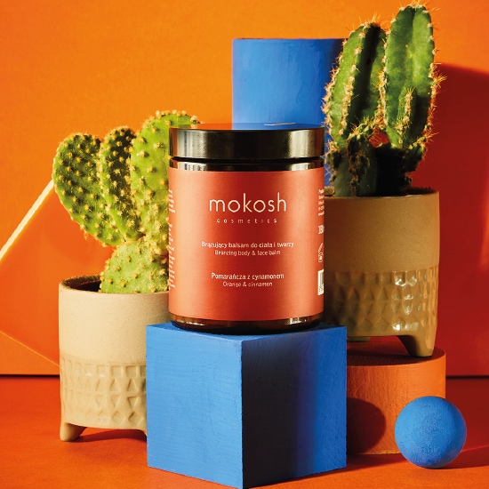 Na pomarańczowym tle, brązowy słoik z czarną nakrętką balsamu do ciała i twarzy Mokosh o zapachu pomarańczy i cynamonu, stoi na niebieskim bloku obok kaktusów w doniczkach.