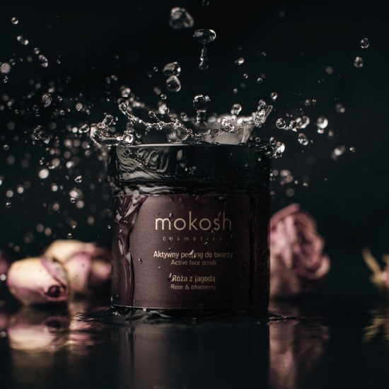 Na ciemnym tle słoik z peelingiem do twarzy Mokosh, otoczony kroplami wody, z etykietą informującą o aktywnym peelingu z róży i jagód.