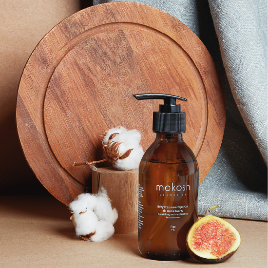 Brązowa butelka żelu do mycia twarzy Mokosh z dozownikiem na drewnianej tacy, obok leżą figi i kwiaty bawełny. Żel odżywczo-nawilżający do mycia twarzy. Odżywia i nawilża skórę.