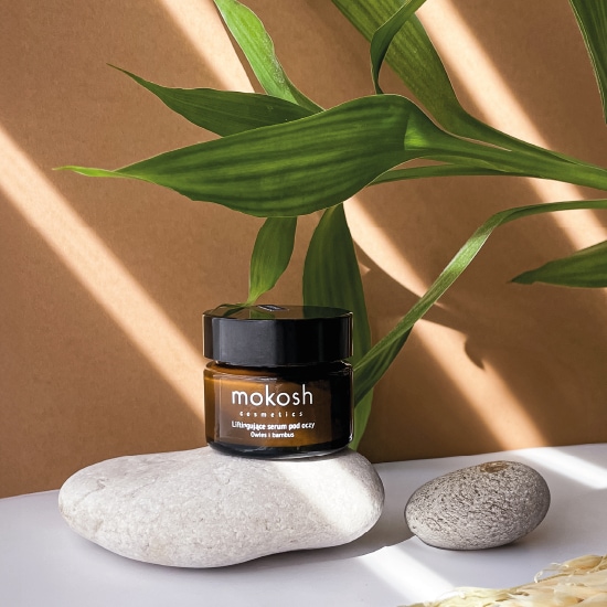 Na pierwszym planie słoiczek z ciemnego szkła z czarną nakrętką, zawierający liftingujące serum pod oczy marki Mokosh. Słoiczek stoi na białym kamieniu, a w tle widać zielone liście bambusa.