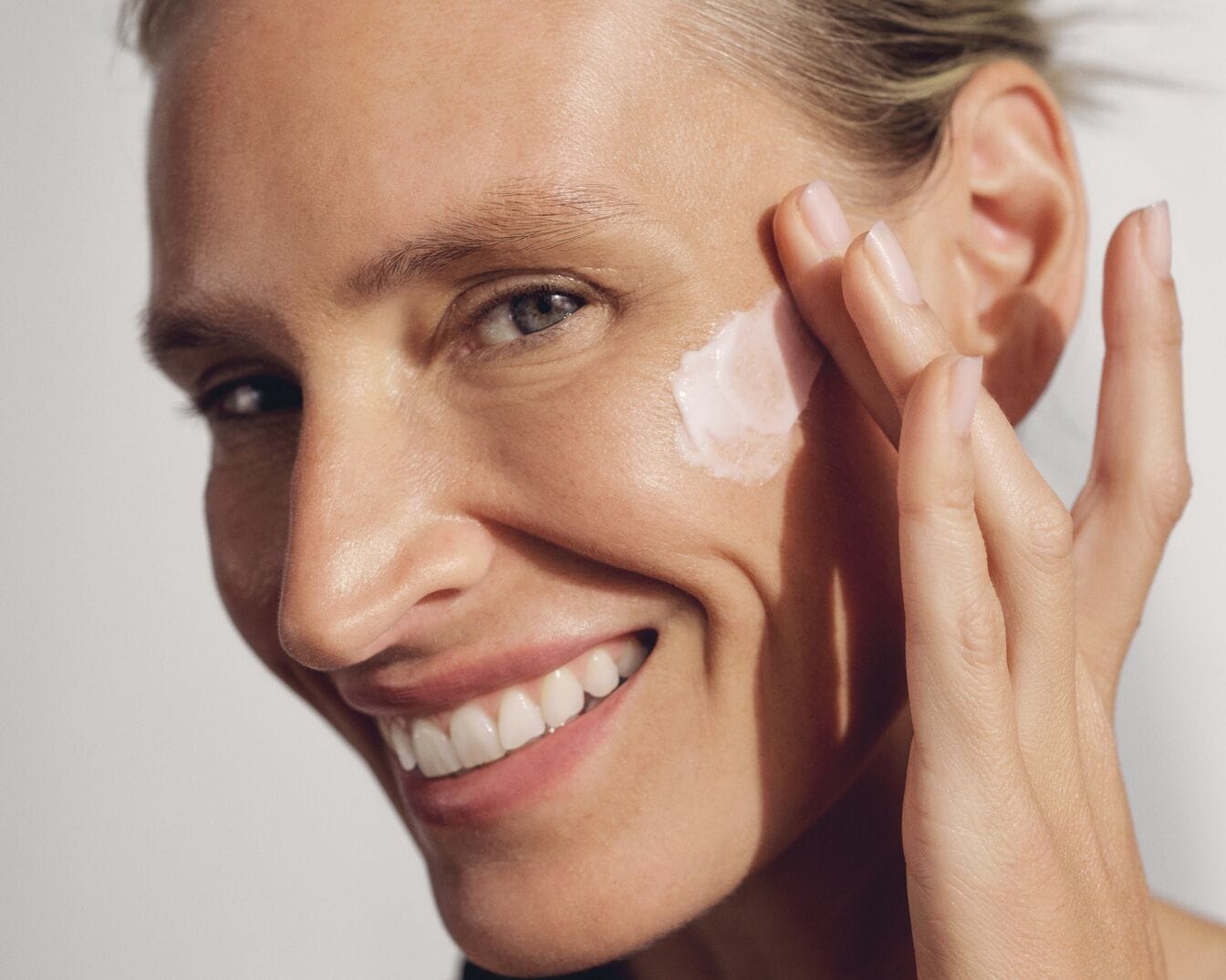 Nahaufnahme einer lächelnden Frau, die eine helle Creme mit den Fingern auf ihre Wange aufträgt, um ihre Haut zu pflegen.