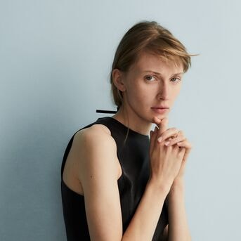 Eine Frau mit blonden, kinnlangen Haaren und einem ärmellosen schwarzen Oberteil posiert vor einem hellblauen Hintergrund.