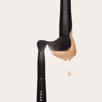 Du juodi makiažo šepetėliai su smėlio spalvos skysta pudra ant šerelių ir lašeliu, krintančiu žemyn.