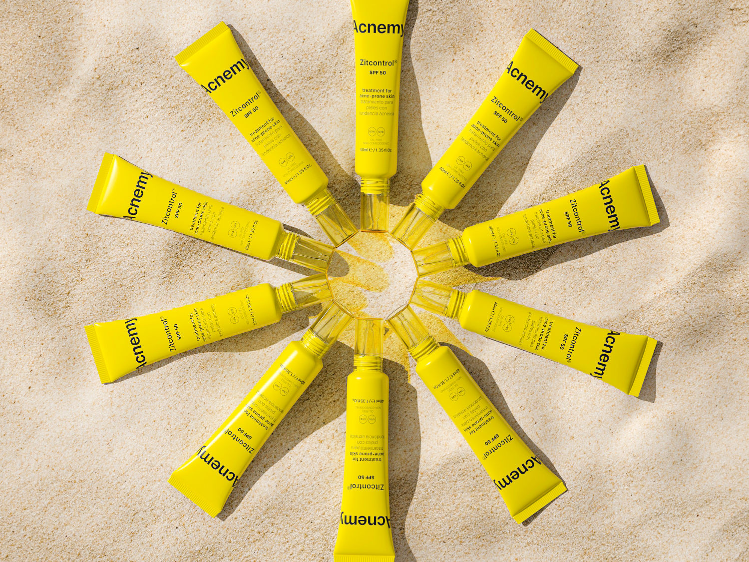 Gelbe Tuben Acnemy Zitcontrol® SPF 50, angeordnet in einem Kreis auf sandfarbenem Untergrund, bieten Behandlung für zu Akne neigende Haut und Schutz vor Sonneneinstrahlung.