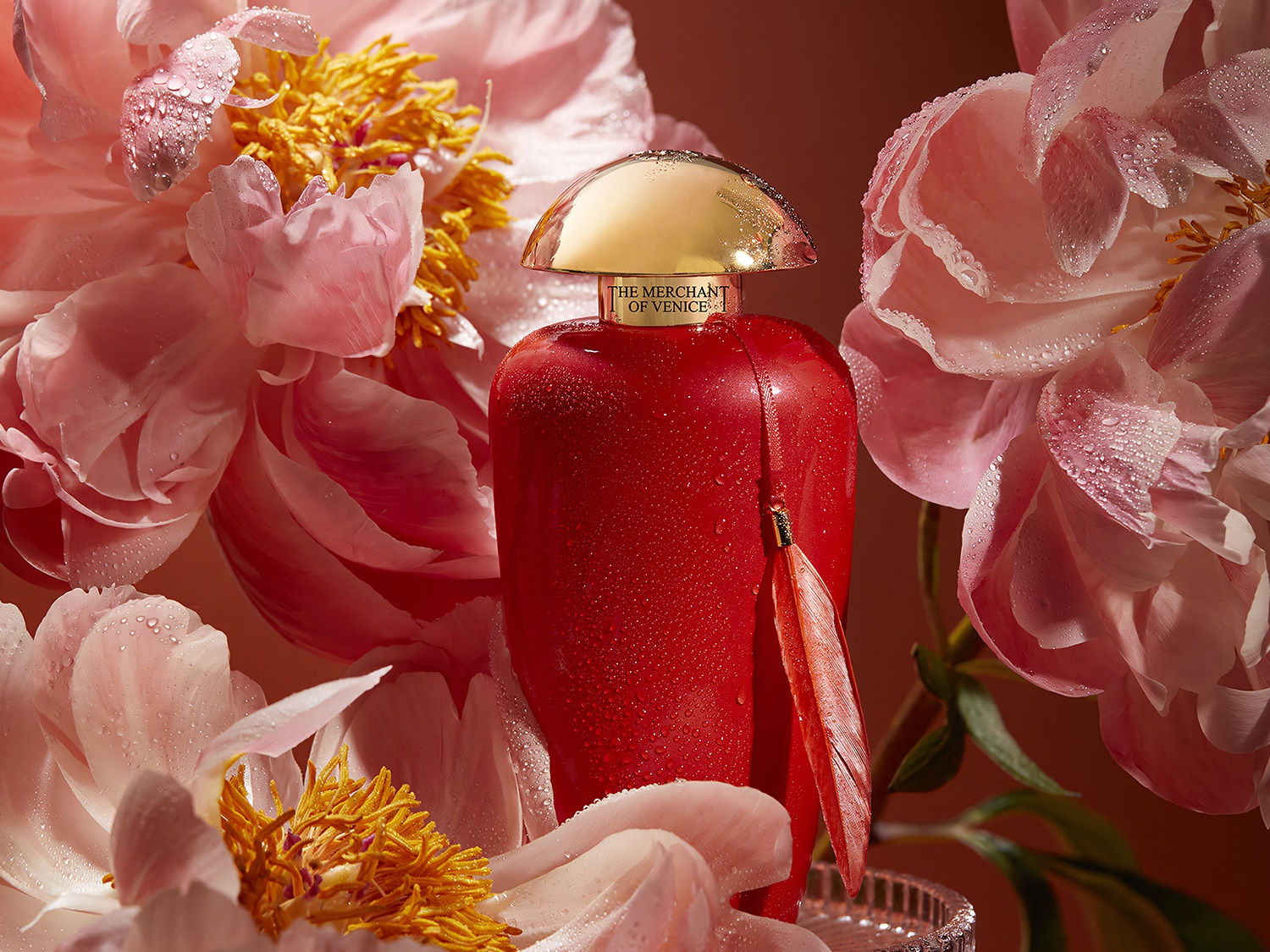 Czerwona butelka perfum The Merchant of Venice z złotą nakrętką, otoczona różowymi kwiatami piwonii z kroplami wody, tworzy luksusową i świeżą kompozycję.
