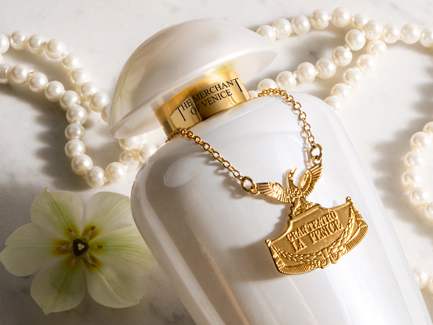 Biały flakon perfum The Merchant of Venice ozdobiony złotym łańcuszkiem z zawieszką i perłami, otoczony białym kwiatem, tworzy luksusową kompozycję.