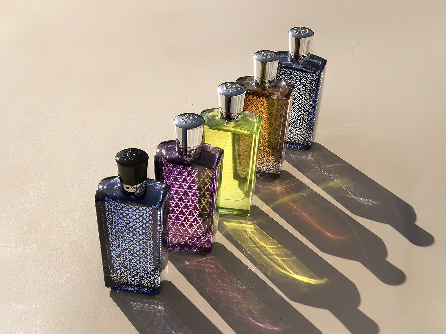 Kolekcja flakonów perfum The Merchant of Venice w różnych kolorach, od ciemnoniebieskiego po jasnozielony, ustawionych w rzędzie na beżowym tle, tworzących długie cienie.
