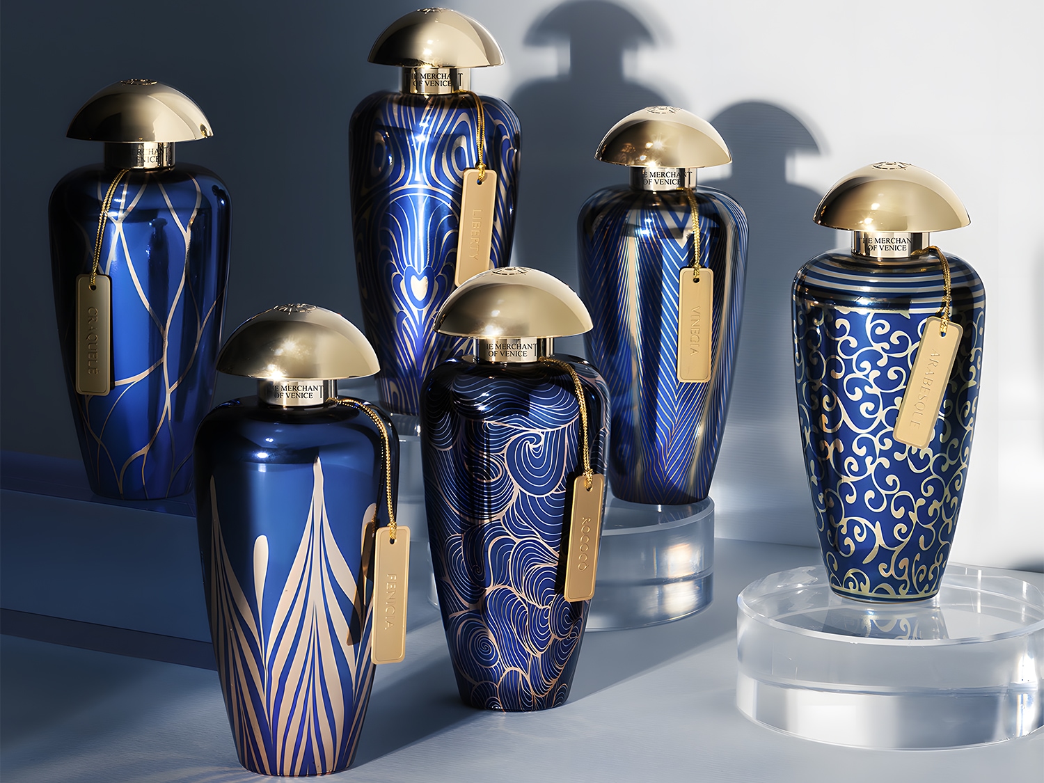 Na zdjęciu znajduje się sześć flakonów perfum The Merchant of Venice w odcieniach granatu i złota, każdy z unikalnym wzorem i złotą zawieszką z nazwą.