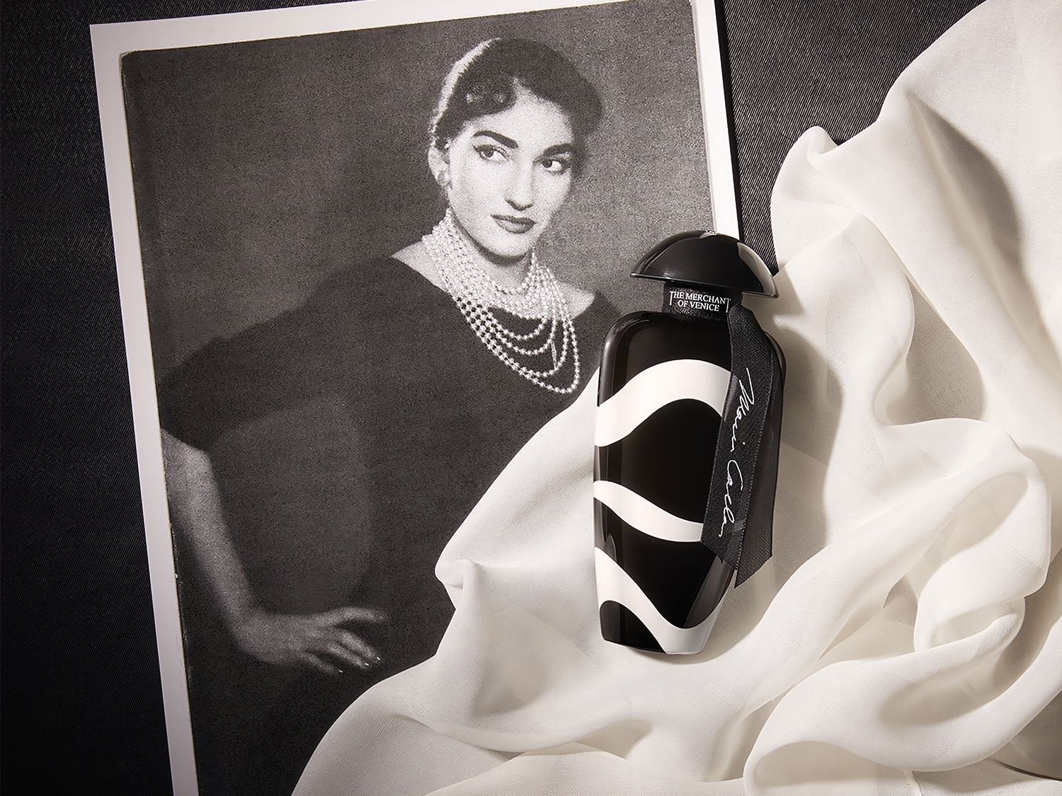 Czarno-biała butelka perfum The Merchant of Venice Maria Callas stoi na białej, zwiewnej tkaninie obok czarno-białego zdjęcia kobiety w sukni i perłach.