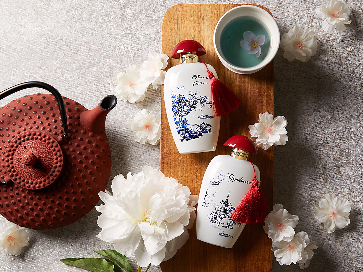 Dwa białe flakony perfum z czerwonymi korkami i frędzlami, ozdobione niebieskimi motywami, leżą na drewnianej desce obok czerwonego czajnika i filiżanki z kwiatem.