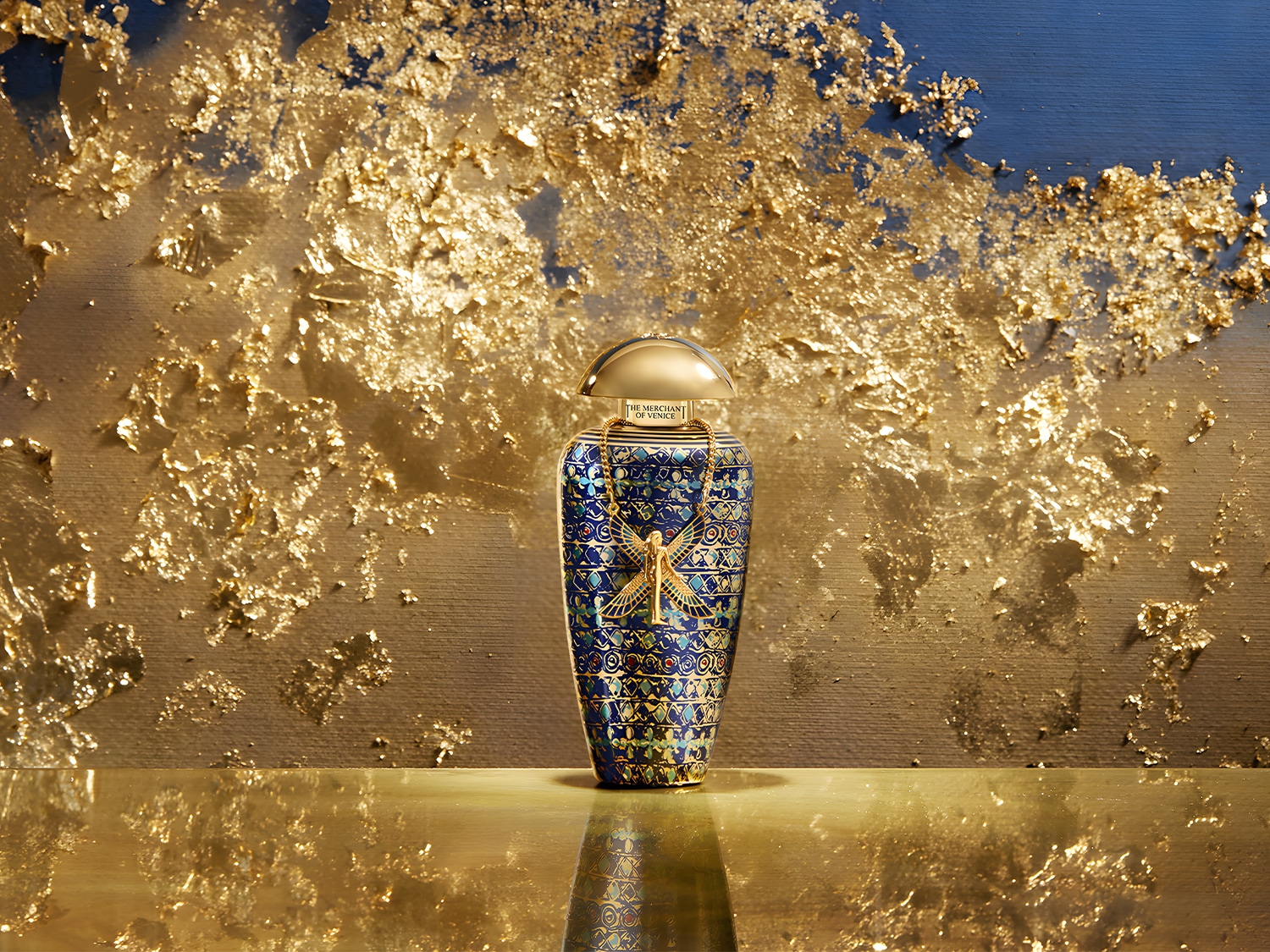 Butelka perfum The Merchant of Venice o pojemności 100 ml, z niebieskimi wzorami i złotymi akcentami, stoi na złotym blacie na tle ściany z efektem złota.