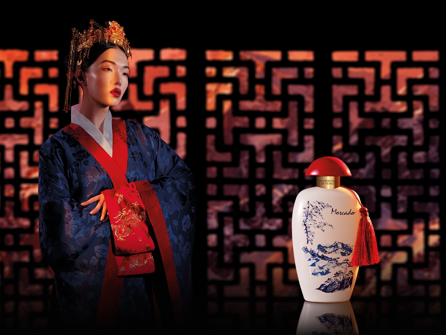 Na tle wzorzystej ściany stoi modelka w tradycyjnym, niebieskim stroju z czerwonymi akcentami i złotą ozdobą na głowie, obok flakon perfum The Merchant of Venice z czerwoną zatyczką i grafiką krajobrazu.