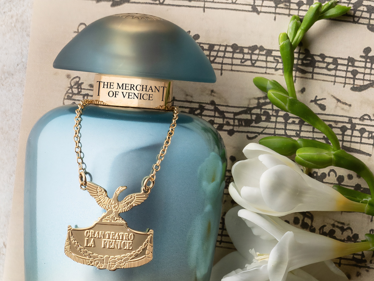 Błękitny flakon perfum The Merchant of Venice z złotym łańcuszkiem i zawieszką Gran Teatro La Fenice, leżący na tle nut i białych kwiatów.