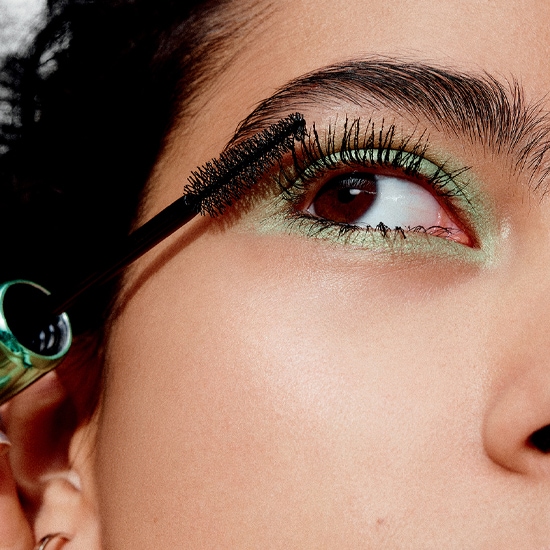 Nahaufnahme einer Person, die mit einer schwarzen Mascara ihre oberen Wimpern tuscht, umrahmt von einem hellgrünen Lidschatten.