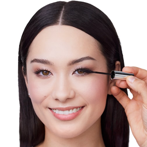 Nahaufnahme einer lächelnden Frau, die sich mit einer silbernen Bürste schwarze Wimperntusche auf die Wimpern aufträgt.