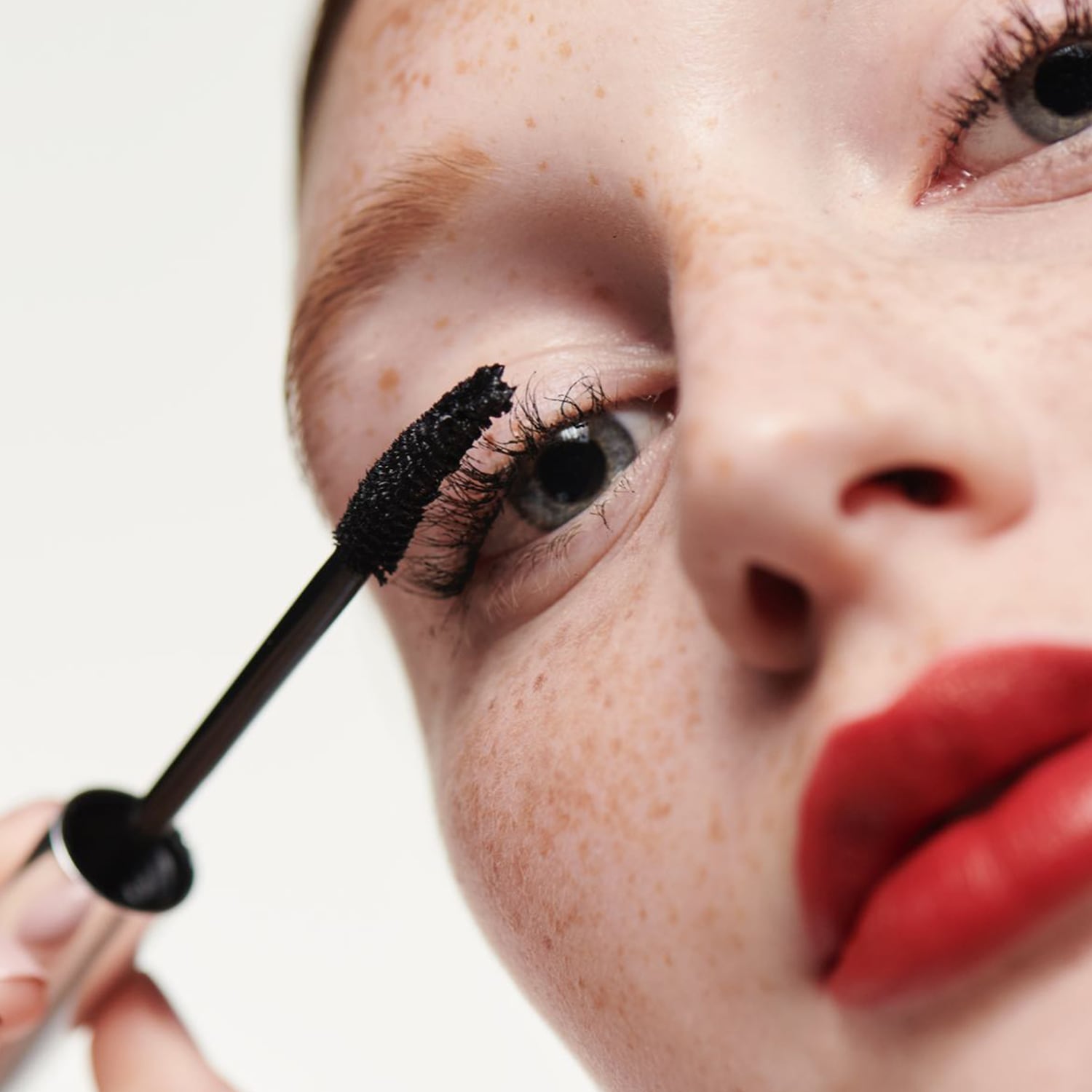 Nahaufnahme einer Frau mit Sommersprossen, die sich mit einer schwarzen Mascara die Wimpern tuscht und roten Lippenstift trägt.