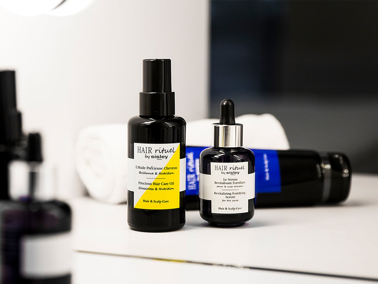 Das Bild zeigt zwei schwarze Flaschen von Hair Rituel by Sisley: Ein Haaröl für Glanz und Pflege und ein revitalisierendes Serum für die Kopfhaut.