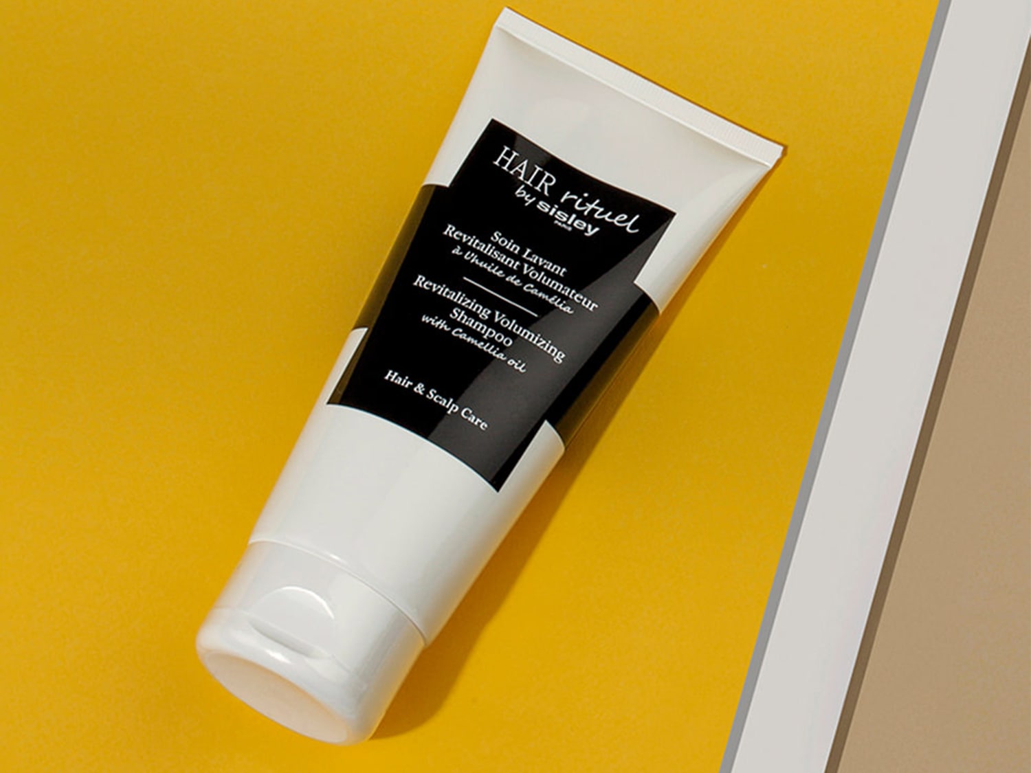 Die weiße Tube Hair Rituel by Sisley Revitalizing Volumizing Shampoo mit Camellia-Öl auf gelbem Hintergrund verspricht Volumen und Pflege für Haar und Kopfhaut.
