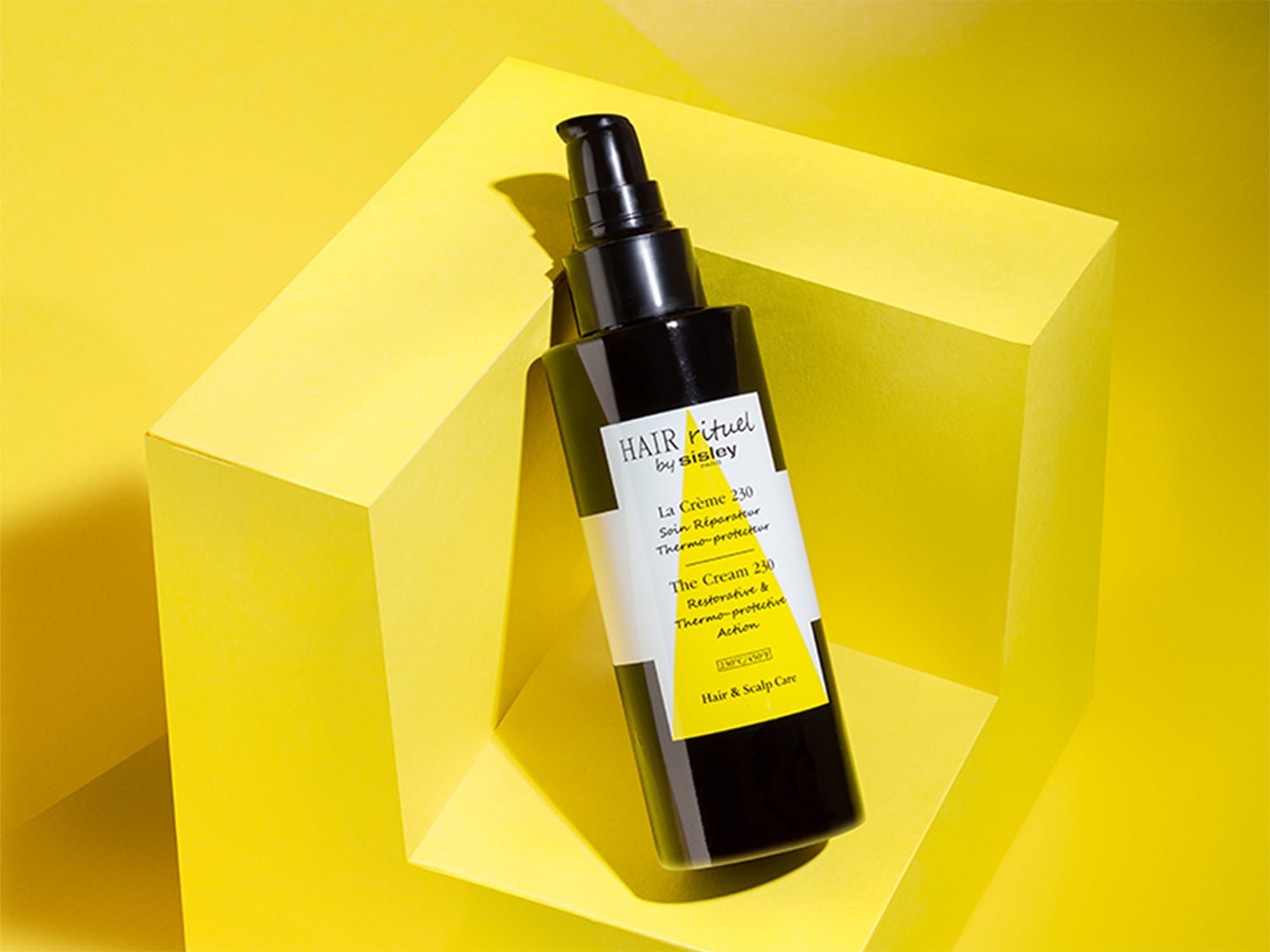 Schwarze Flasche Hair Rituel by Sisley La Crème 230, eine restaurative und thermisch schützende Creme für Haar und Kopfhaut, vor einem gelben geometrischen Hintergrund.