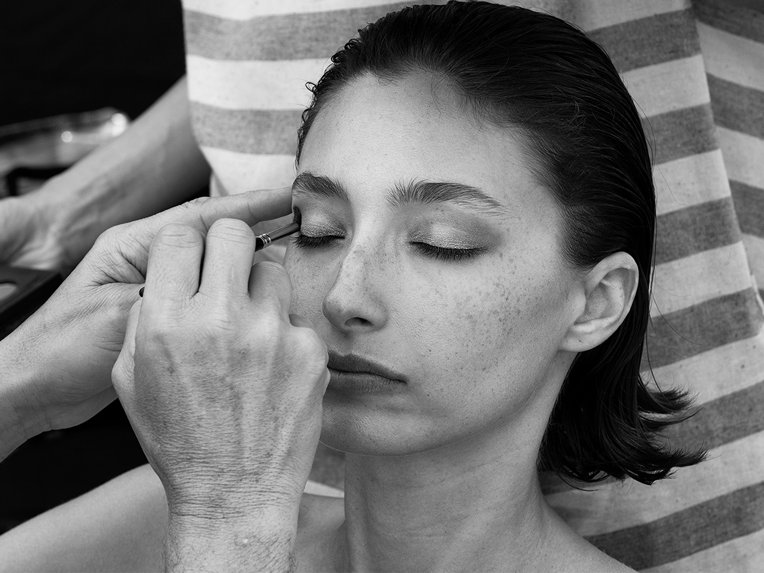 Primo piano in bianco e nero di una modella con gli occhi chiusi mentre un truccatore le applica l'eyeliner per migliorare il suo sguardo.