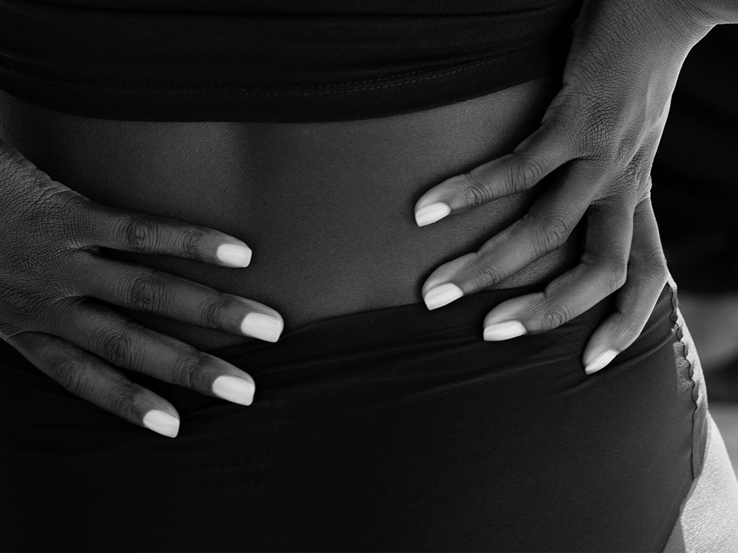 In un'immagine in bianco e nero, una persona con la pelle scura indossa un top e una parte inferiore neri, con le mani appoggiate sui fianchi e le unghie dipinte di bianco.