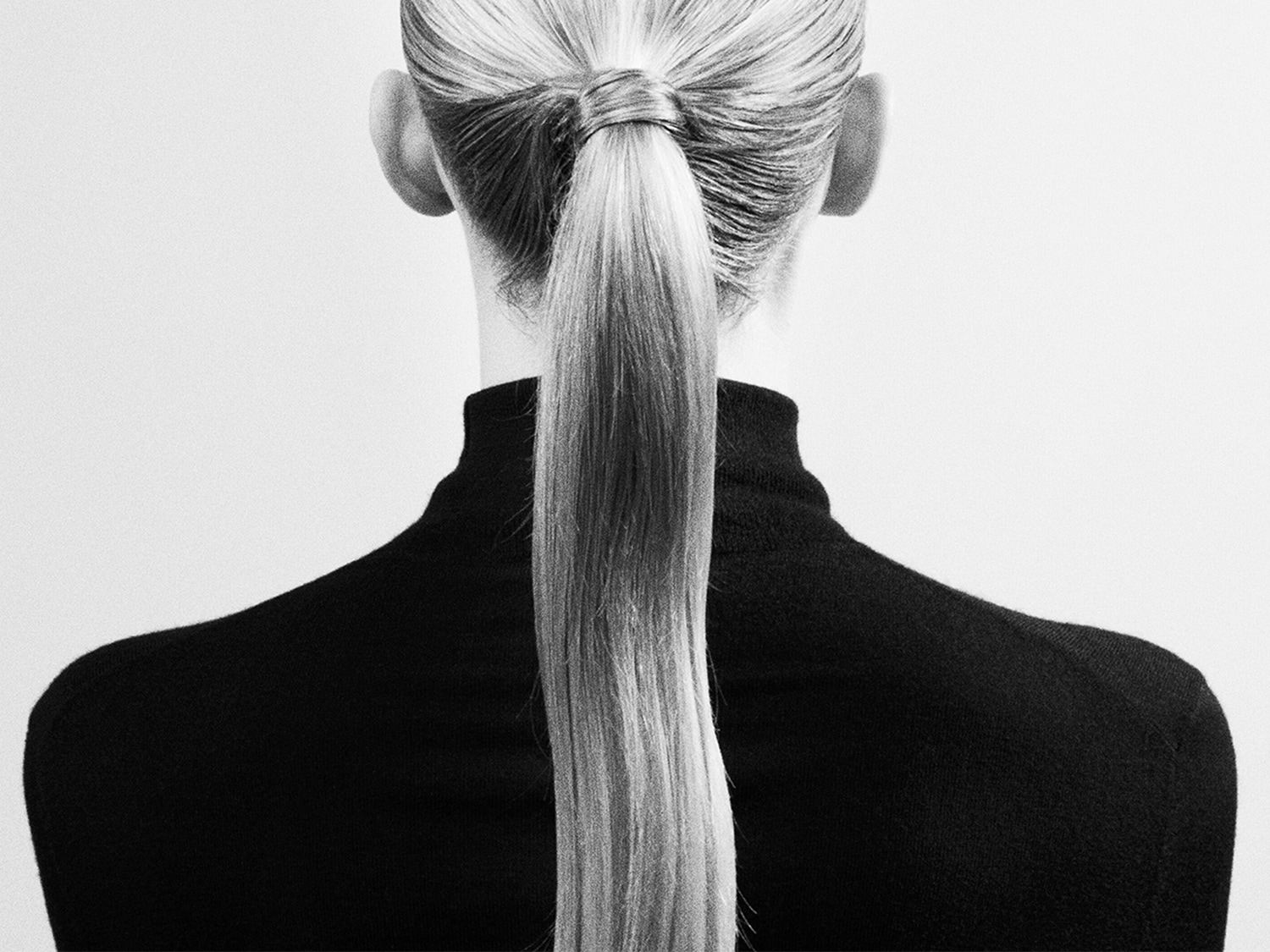 Vista posteriore di una donna con capelli biondi raccolti in una coda di cavallo, indossa un dolcevita nero, creando un contrasto elegante e minimalista.