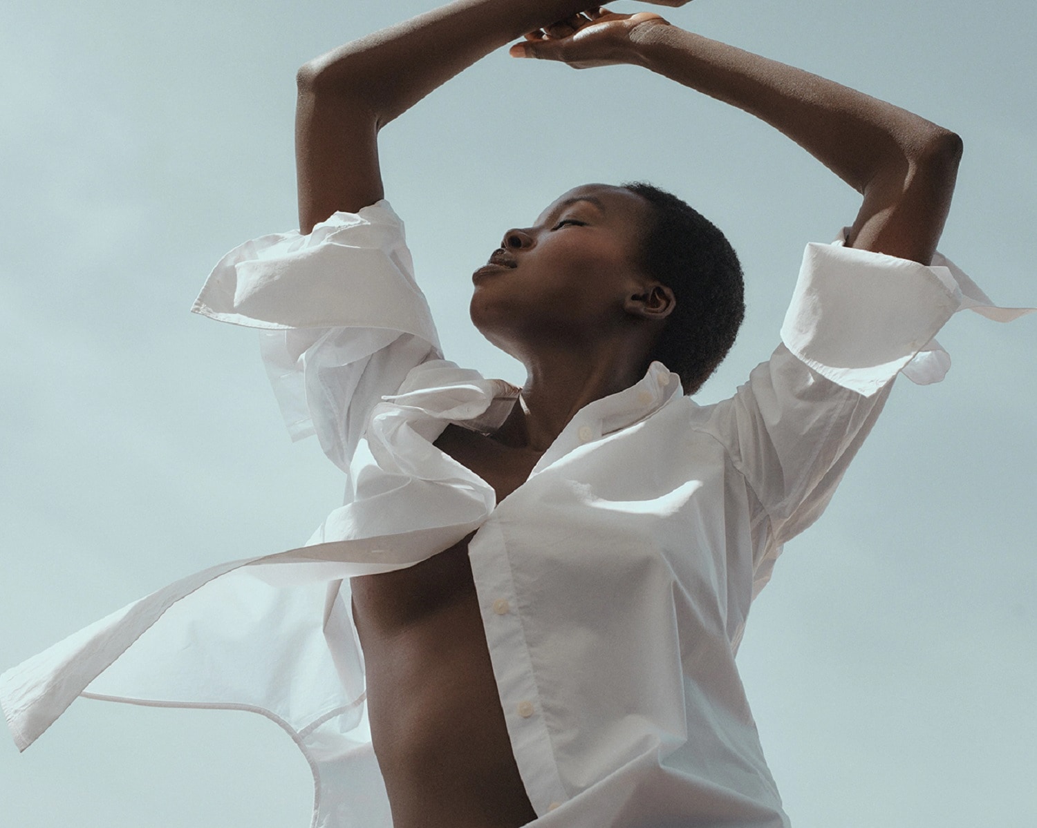 Eine Frau mit dunkler Haut und kurzen, dunklen Haaren trägt ein weißes Hemd und streckt die Arme in den Himmel.
