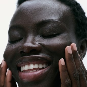 Nahaufnahme einer lächelnden, dunkelhäutigen Frau mit geschlossenen Augen, die ihre Wangen berührt und eine strahlende, glänzende Haut zeigt.