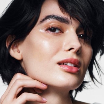 Nahaufnahme einer Frau mit glänzender Haut und glänzenden Lippen, die ihre Wange berührt, was einen frischen und strahlenden Teint zeigt.