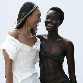 Zwei Frauen unterschiedlicher Hautfarbe umarmen sich und lachen, die eine in Weiß, die andere in dunkler Bademode.