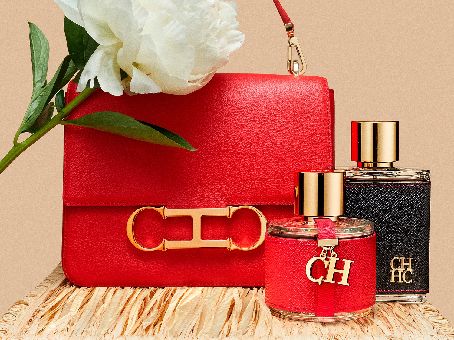 Bolso rojo elegante junto a dos frascos de perfume CH con detalles dorados y una peonía blanca, sobre una superficie texturizada.