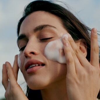 Eine Frau mit geschlossenen Augen massiert sanft weißen Schaum auf ihr Gesicht, um ihre Haut zu reinigen und zu pflegen.