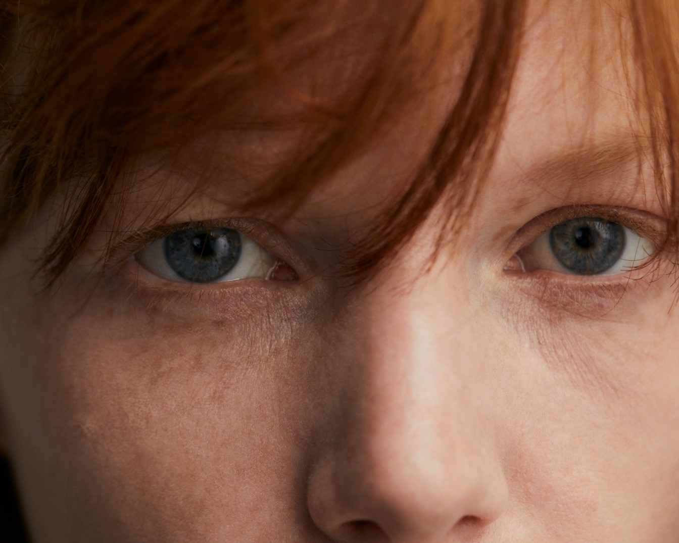 Zbliżenie na twarz kobiety o jasnej karnacji, z rudymi włosami opadającymi na czoło i intensywnie niebieskimi oczami.