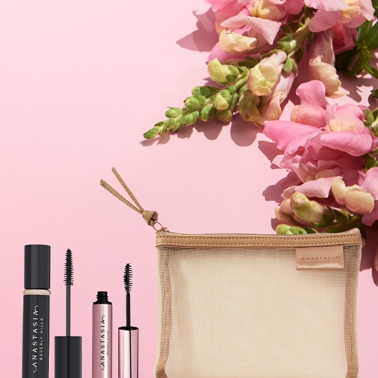 Na różowym tle leżą dwa tusze do rzęs Anastasia Beverly Hills w czarnym i różowym opakowaniu, obok nich znajduje się kosmetyczka z siateczki.