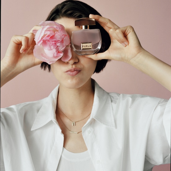 Młoda kobieta w białej koszuli trzyma flakon perfum Furla i różowy kwiat przy oczach, na różowym tle.