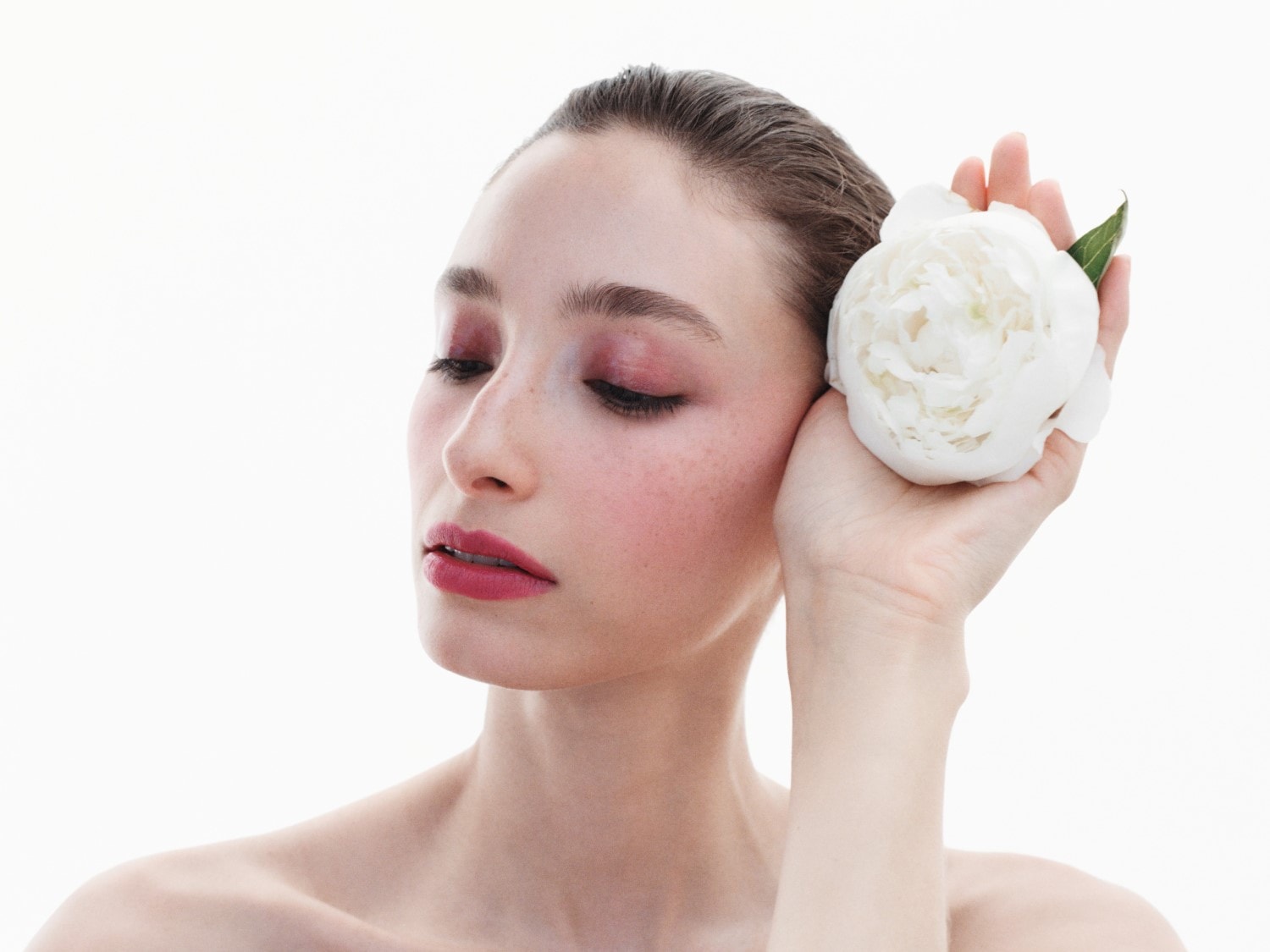 Eine Frau mit rosa Rouge und Lippenstift hält eine weiße Pfingstrose an ihr Gesicht.