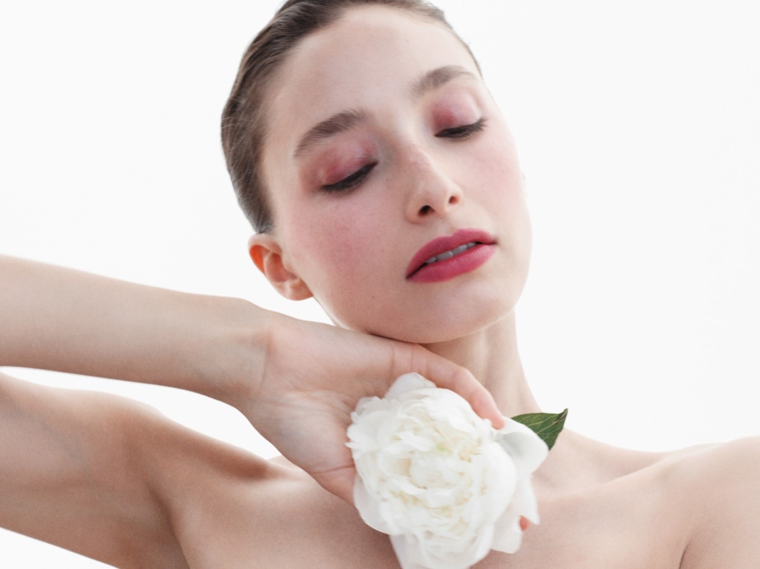 Eine Frau mit geschlossenen Augen, zarter rosa Schminke und einem weißen Pfingstrosenblüte in der Hand.