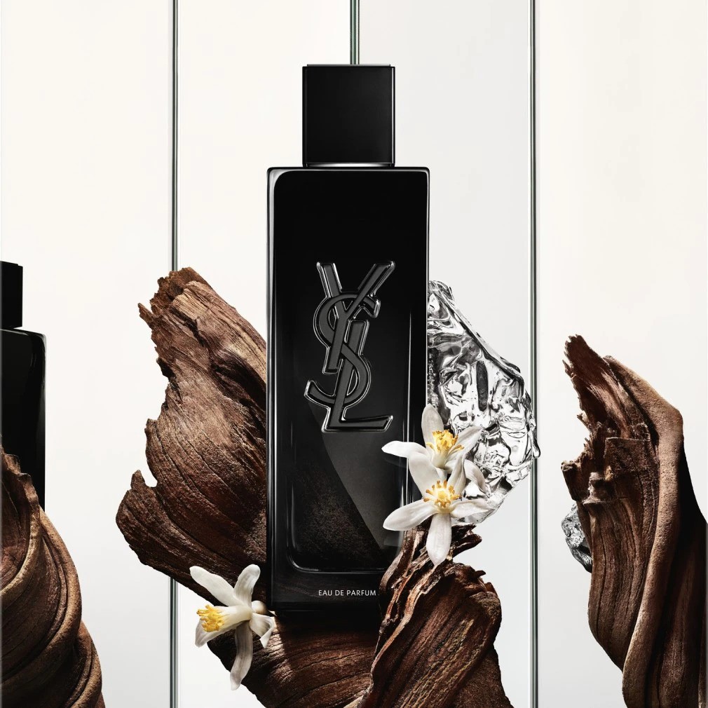 Zwarte YSL Eau de Parfum fles met zilveren logo, geplaatst tussen stukken hout, een stuk zilverkleurige folie en witte bloemen.