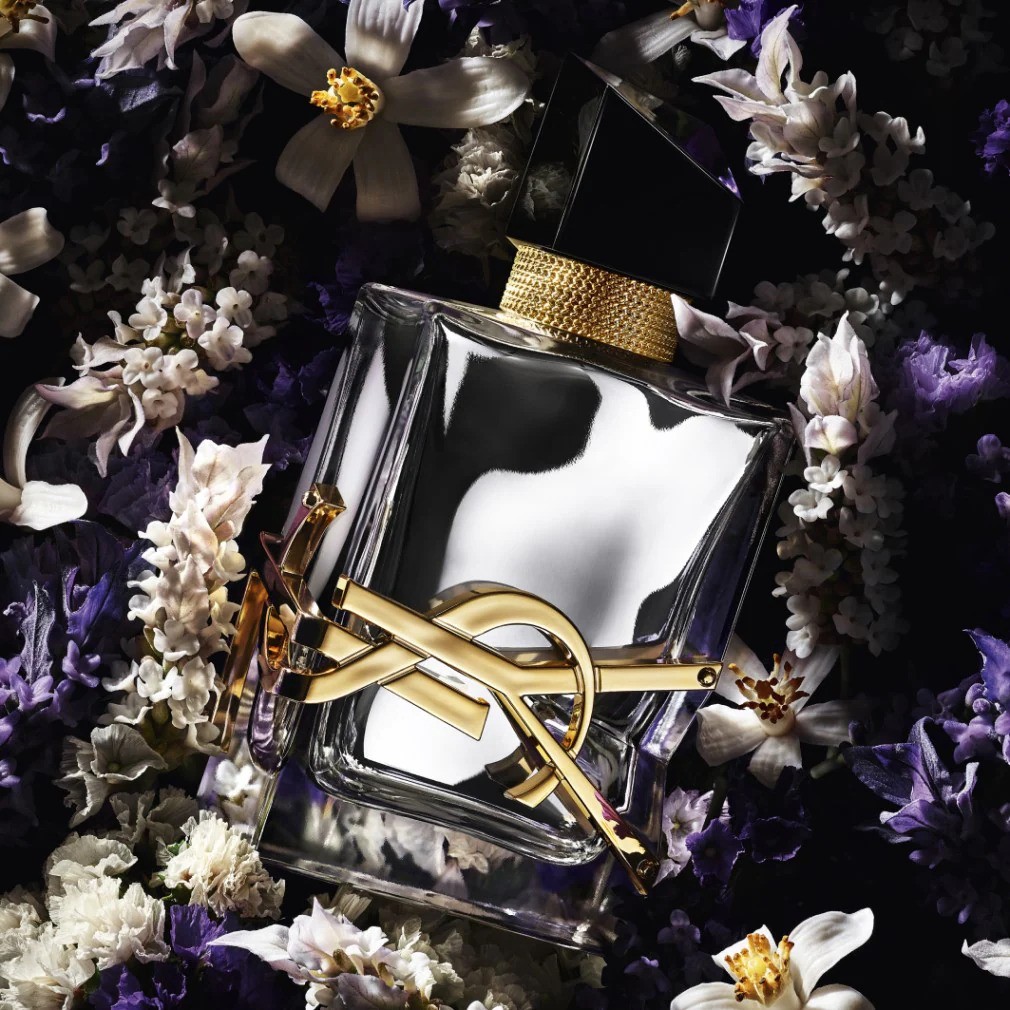 Een heldere glazen parfumfles met een gouden YSL-logo ligt te midden van een bed van witte en paarse bloemen.