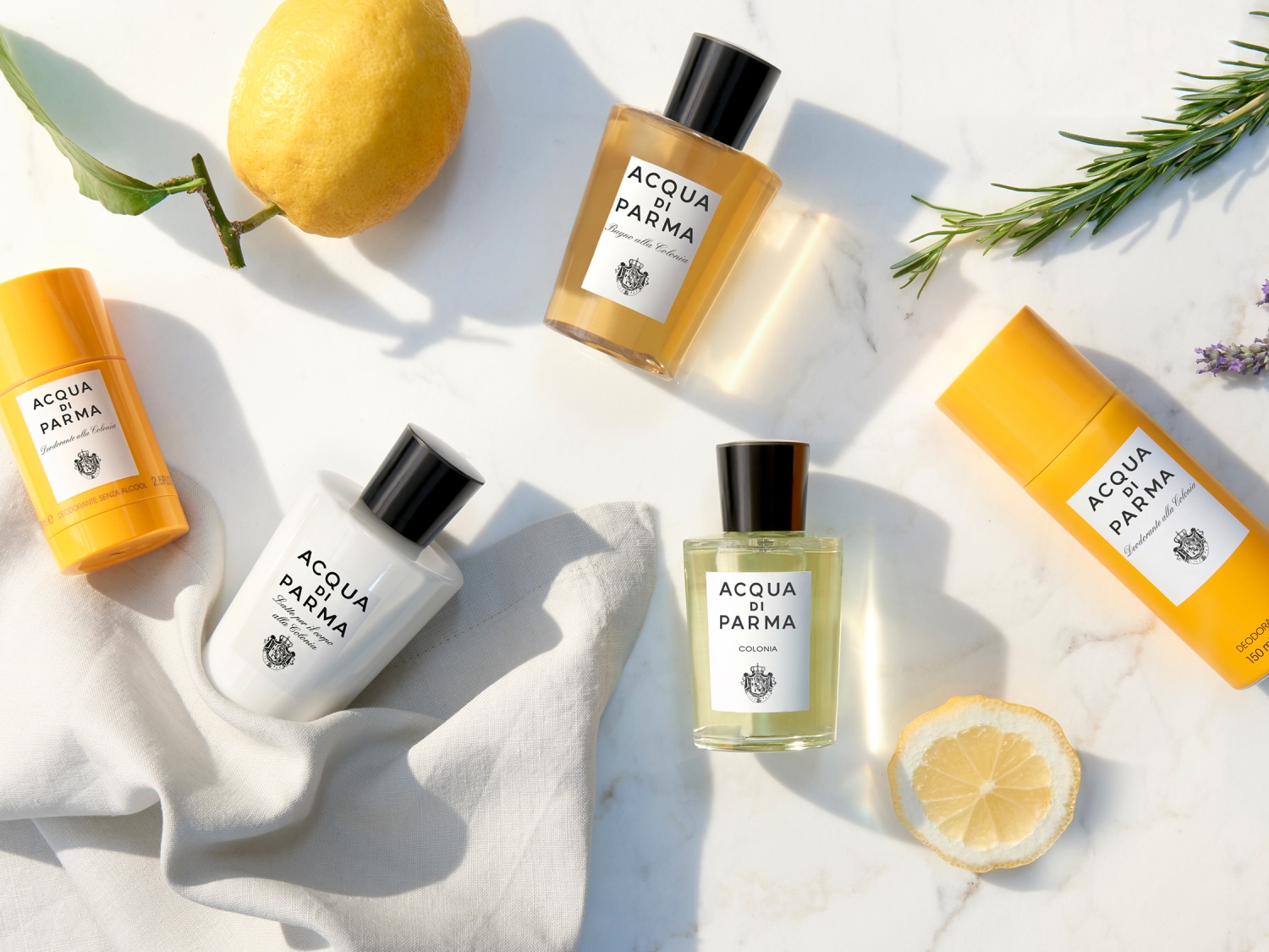Na marmurowym tle leżą produkty Acqua di Parma: żółty dezodorant w sztyfcie, biały balsam do ciała, bursztynowy żel pod prysznic, woda kolońska w przezroczystej butelce, żółty dezodorant w sprayu, cytryna, plasterek cytryny, gałązka rozmarynu i lawendy.
