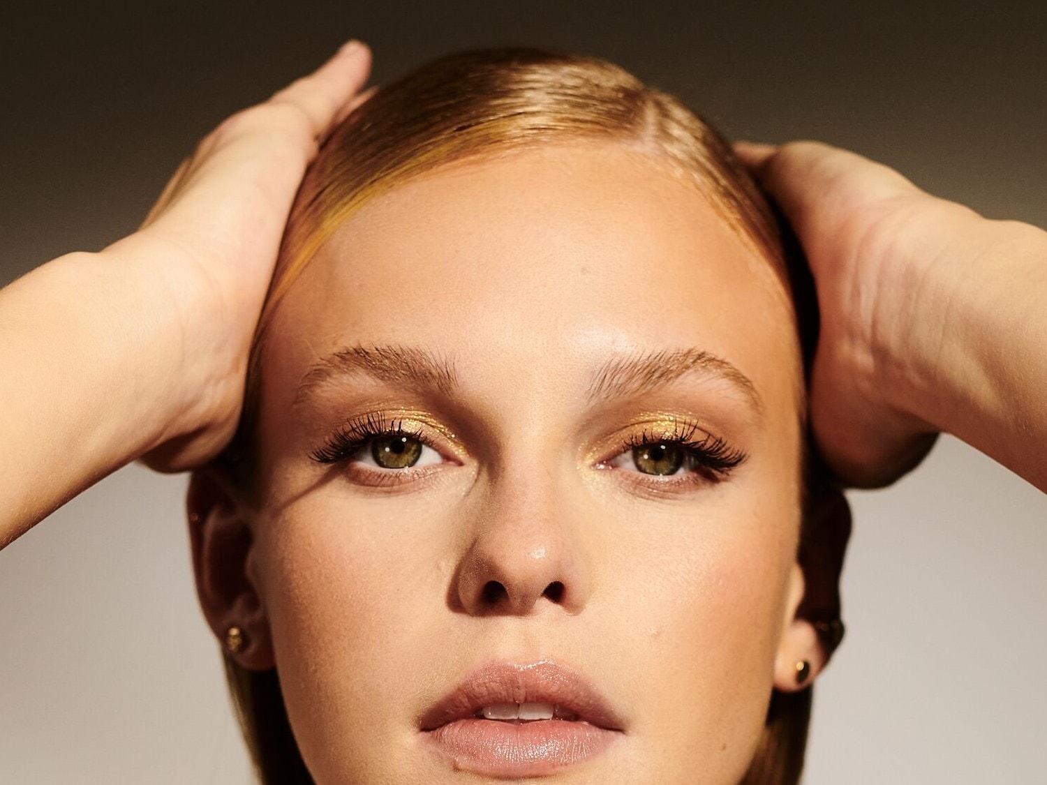 Nahaufnahme eines Gesichtes mit goldenem Lidschatten und glänzenden Lippen, die Hände berühren das Haar.