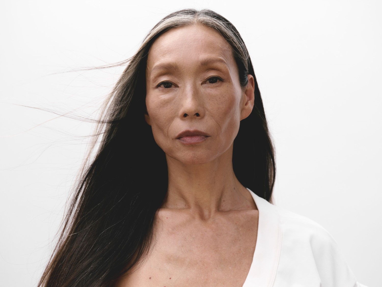Porträt einer Frau mit langen, glatten, schwarz-grauen Haaren, die einen weißen Blazer trägt und einen ruhigen, nachdenklichen Ausdruck hat.