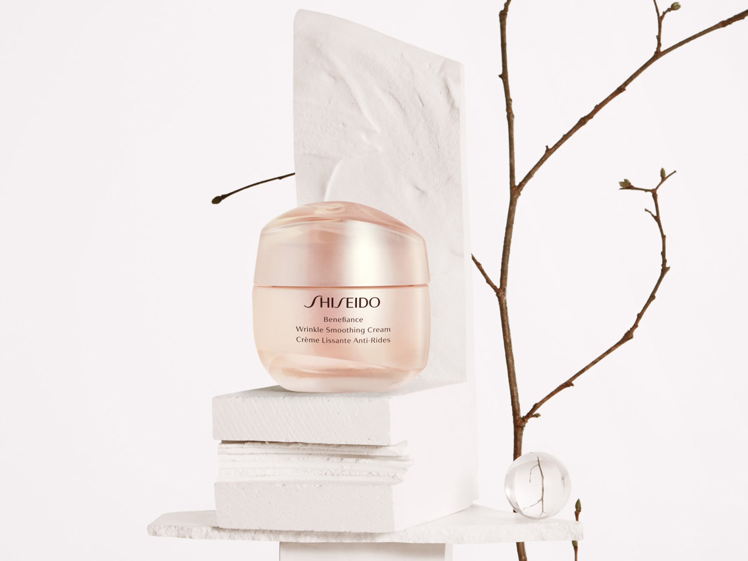 Il vasetto color pesca di Shiseido Benefiance Wrinkle Smoothing Cream è in mostra su blocchi bianchi, accanto a un ramo spoglio e una sfera di vetro, per una presentazione elegante.