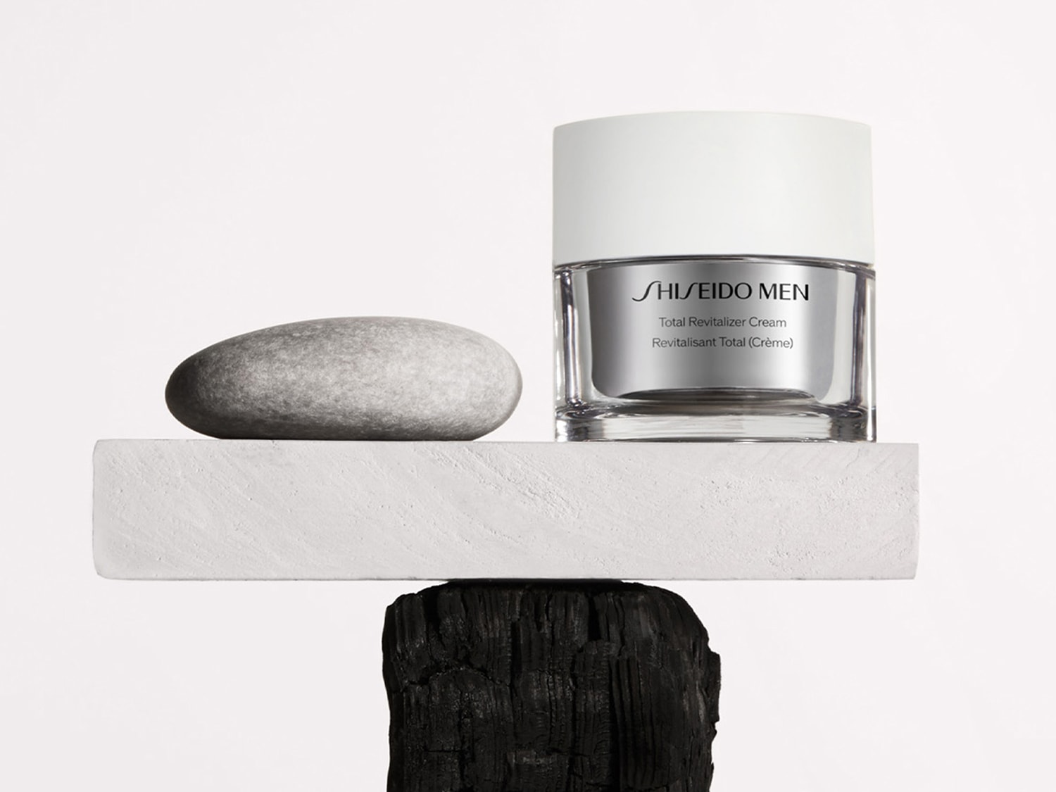 Crema rivitalizzante totale Shiseido Men in confezione argento e bianca, appoggiata su una piattaforma bianca sopra un blocco di carbone, con una pietra grigia liscia accanto.