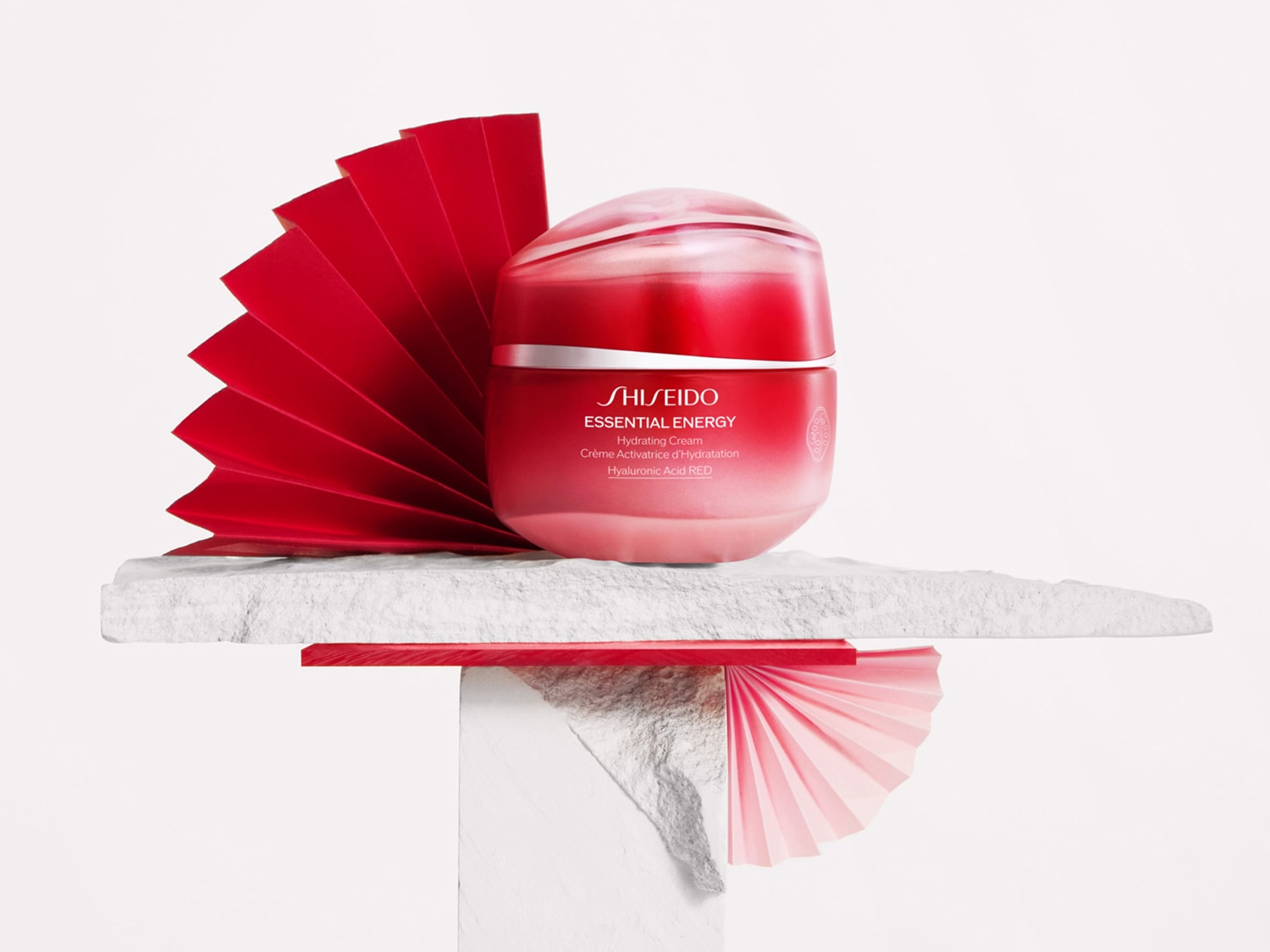 Vaso rosso di crema idratante Shiseido Essential Energy su una piattaforma bianca con decorazioni a ventaglio rosse, per un'idratazione intensa e una pelle radiosa.